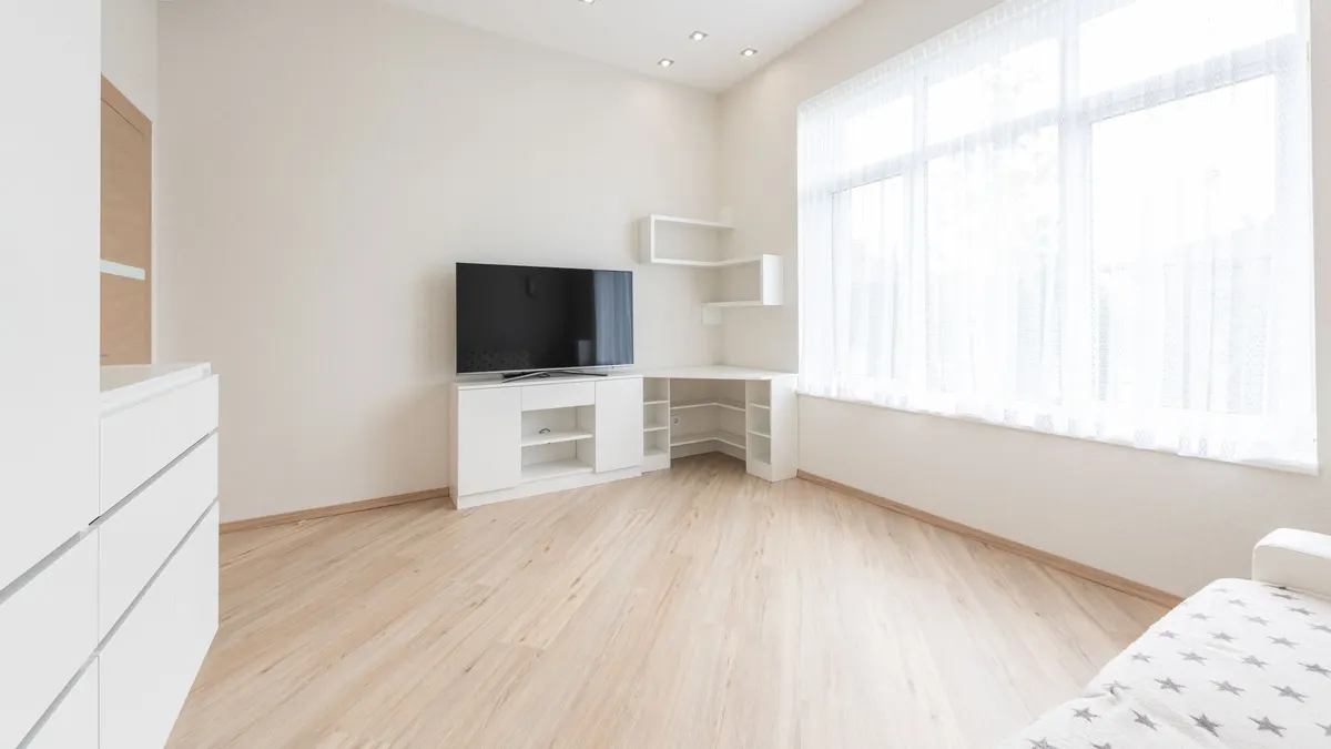 A spacious room with light plank flooring, a low cupboard and a television