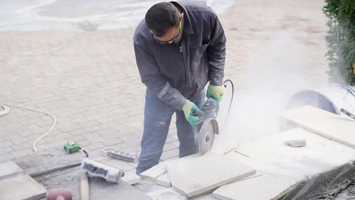 A construction worker using a power saw to cut stone outdoors amid dust