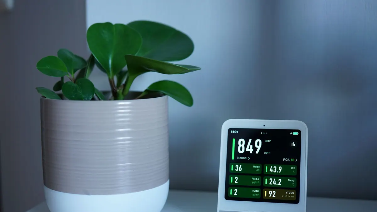 A digital air quality monitor on a shelf beside a potted plant