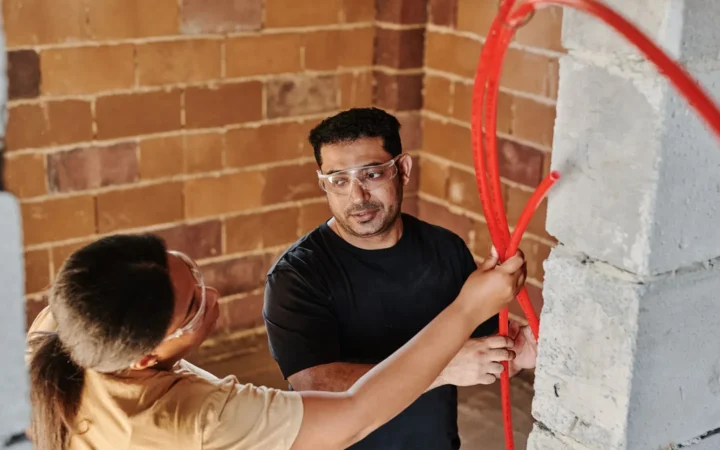 Two people fitting a red pipe against a brick wall indoors