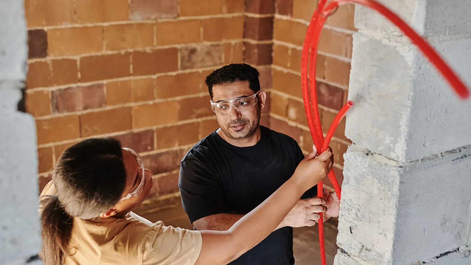 Two people fitting a red pipe against a brick wall indoors
